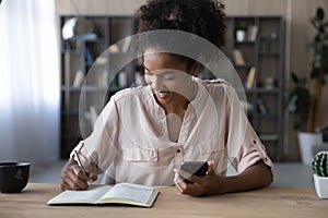 Smiling African American woman holding phone, taking notes in notebook