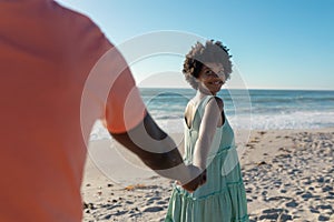 Smiling african american woman looking back while holding hand of boyfriend at beach