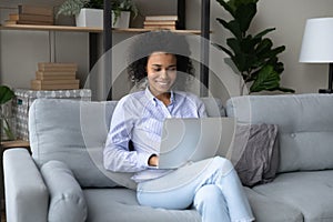 Smiling African American woman using laptop, relaxing on cozy couch