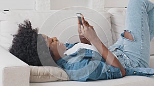 Smiling African American woman using smartphone dating app looking swiping on screen, happy mixed race lazy young girl