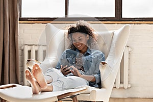 Smiling African American woman using smartphone, relaxing in armchair