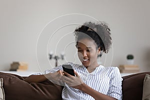 Smiling African American young woman using smartphone, sitting on couch