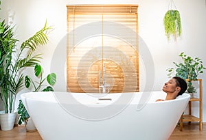 Smiling African attractive woman wearing headphones relaxing and dancing in foam bath in bathroom.