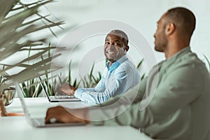 Smiling african businessman working on computer sitting in coworking on colleagues background