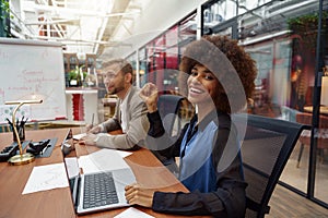 Smiling african businesswoman sitting on his workplace in modern office on colleague background