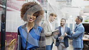 Smiling african businesswoman standing in office and looking at camera with colleagues in background