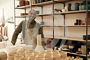 Smiling African ceramist standing by a bench and shelves full of pots