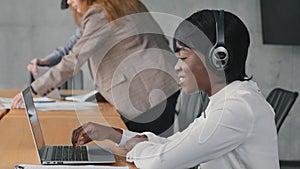 Smiling African female call center agent wear wireless headset using laptop looking at screen consulting customer with