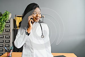 Smiling african female doctor physician holding cell phone talking on mobile at work. Healthcare professional answering
