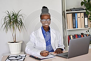 Smiling african female doctor wear glasses looking at camera sit at workplace.