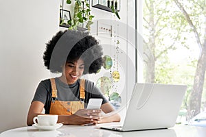 Smiling African hipster teenage girl using smart phone sitting at cafe table.