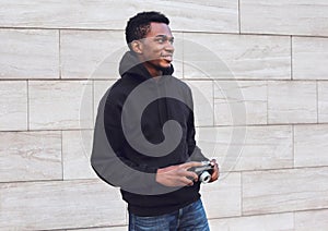 Smiling african man photographer with vintage film camera looking away wearing black hoodie while walking on city street