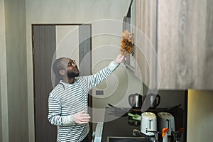 Smiling African man wiping dust off the furniture with a feather duster. Housekeeping concept