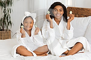 Smiling African Mom And Daughter Sitting In Bathrobes Holding Cucumber Slices