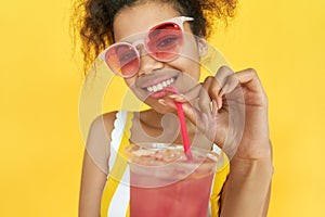 Smiling african teen girl drinking cocktail wear sunglasses isolated on yellow.