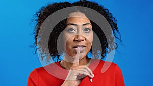 Smiling african woman with curly hair holding finger on her lips over blue background. Gesture of shhh, secret, silence