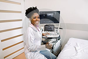 Smiling African woman doctor with ultrasound scanner in hand, working on modern ultrasound scanning machine in light
