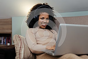 Smiling african woman in headphones using laptop computer while sitting on a couch at home