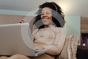 Smiling african woman in headphones using laptop computer while sitting on a couch at home