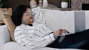 Smiling african woman student girl freelancer user lying on couch using laptop typing message online chat with friends