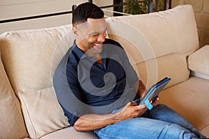Smiling african young man sitting on sofa and using digital tablet at home
