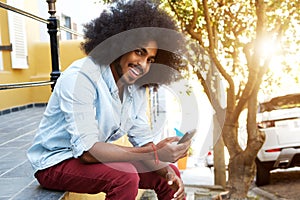 Smiling afro man sitting outside by sidewalk with mobile phone