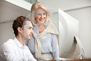 Smiling aged female mentor looking at computer screen helping in