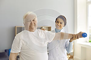 Smiling aged man patient doing physiotherapy exercise with dumbbell under woman chiropractors control