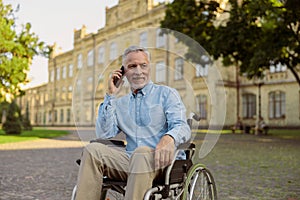 Smiling aged man recovering patient in wheelchair looking away while talking on the phone, spending time alone in the