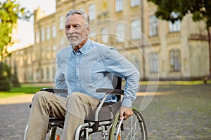 Smiling aged recovering man in wheelchair looking away while spending time alone in the park near hospital