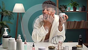 Smiling Arab guy with applying facial moisturizer while holding jar and looking at camera at home. Handsome man applying