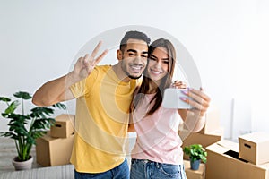 Smiling Arab guy and his Caucasian girlfriend moving to their own home together, taking selfie among carton boxes