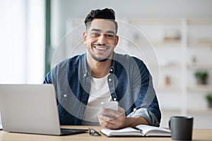 Smiling arab guy using smartphone while sitting at workdesk