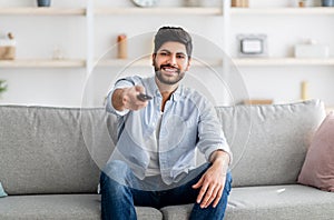 Smiling arab guy watching comedy on TV at home, sitting on sofa in living room, holding TV remote, copy space