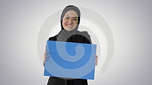 Smiling Arab woman in hijab holding blank blue poster and looking at camera on gradient background.