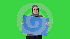 Smiling Arab woman in hijab holding blank blue poster and looking at it on a Green Screen, Chroma Key.