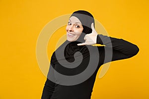 Smiling arabian muslim woman in hijab black clothes doing phone gesture like says call me back isolated on yellow wall