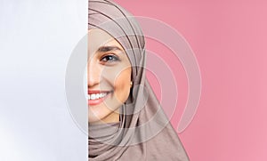 Smiling arabic girl standing behind blank advertisement board, covering half face