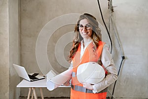 Smiling architect woman in construction site holding blueprints and helmet. Home renovation