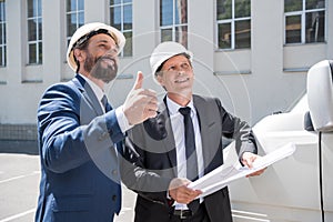 Smiling architects in hardhats holding blueprint and showing thumb up