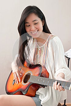 Smiling asian beautiful girl sitting in the music room and playing guitar.