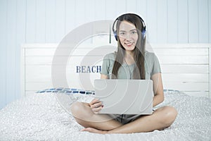 Smiling asian beauty woman sitting on the bed listening music from headphone and playing notebook computer on bed.