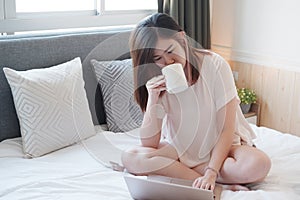 Smiling Asian businesswoman is drinking a cup of coffee and working with laptop in bedroom on holiday. She is online working from