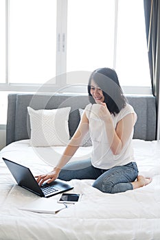 Smiling Asian businesswoman is drinking a cup of coffee and working with laptop in bedroom on holiday. She is online working from