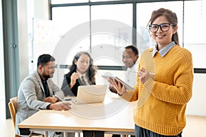 A smiling Asian businesswoman in glasses standing with a tablet in her hand in a meeting room