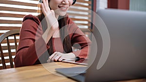 Smiling Asian businesswoman in headset waving hand, using laptop, looking at screen, student learning online