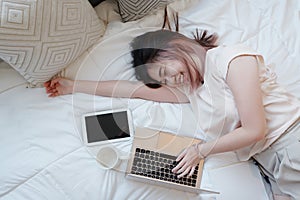 Smiling Asian businesswoman is relaxing and laying with laptop in bedroom on holiday. She is online working from home with