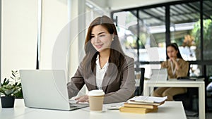 Smiling Asian businesswoman working on her assignment on her laptop at her desk