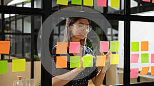 Smiling asian businesswoman write tasks on sticky notes on glass wall