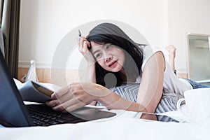 Smiling Asian businesswoman is writing and researching with laptop in bedroom on holiday. She is online working from home with